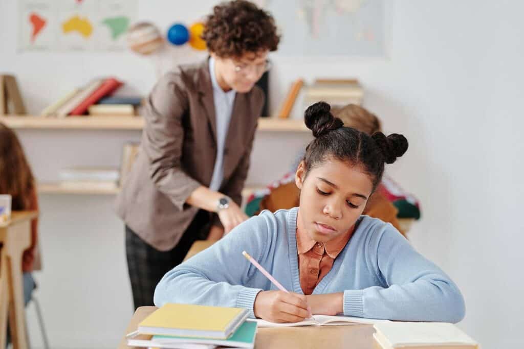 Student frowning while writing on paper with teacher in the background