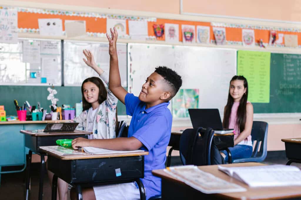 Students in classroom with two raising their hands at the teacher and one looking at the camera.